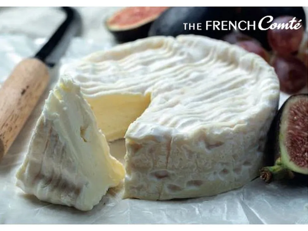 A disc of Camembert cheese, partially cut, with a triangular piece alongside, placed on a white surface with a knife and figs