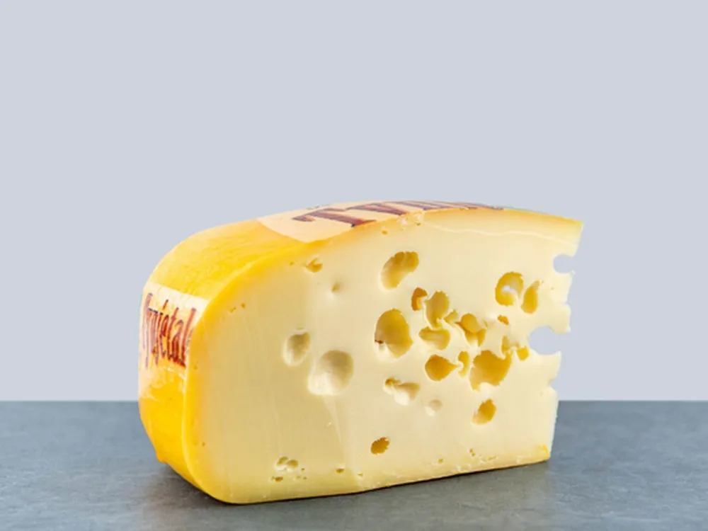 A slice of a Maasdam cheese with a bright yellow rind and a pale yellow interior with eyes (holes)