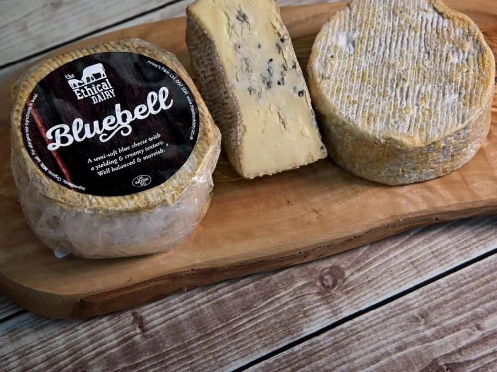 A disc of Bluebell cheese with a label, another without, and a half-cut piece revealing blue veining and a smooth interior, resting on a wooden board