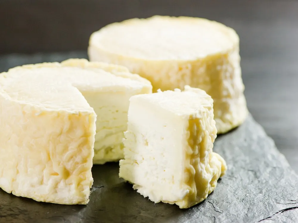 A partially cut wheel of Manchester cheese and a whole wheel, both showing a luscious cream line beneath a thin, wrinkly rind, rest on a black surface