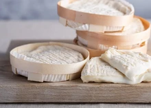 Pérail de Brebis (Pérail) cheese discs and wedges are stacked, displaying their wrinkled pale rind and smooth interior, all set on a wooden surface.