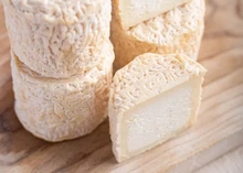 Small discs of Petit Blaja cheese, some stacked and others cut in half to reveal their creamy, soft texture and wrinkled yellow-orange rind, are arranged on a wooden surface