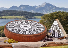 A wheel of Le Gruyère AOP cheese, accompanied by a triangular slice with a label on top, placed on a wooden board with a scenic backdrop of mountains and a river