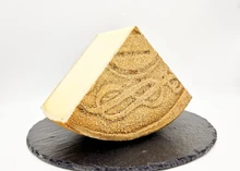 A slice of Alisia-Victoria cheese rests on a round marble surface, showcasing its golden-yellow rind and smooth interior