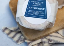 A disc of Baby Katherine cheese, wrapped in white paper with a label on top, rests on a wooden cutting board and a linen napkin