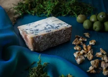 A wedge of Bayley Hazen Blue cheese, streaked with blue veins and encased in a dark brown rind, is elegantly arranged alongside fresh herbs, nuts, and olives, all resting on a rich, dark blue velvet cloth
