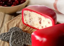 A half-cut small disc of Bouncing Berry cheese showcases its interior infused with flecks of cranberries, accompanied by crackers and fresh cranberries, all arranged on a wooden surface