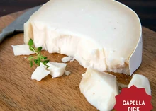 A partially sliced wheel of L'Amuse Brabander Goat Gouda, with chunks scattered on a wooden board, accompanied by fresh herbs and a cutting knife