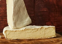 Triangular slices of Brick Lane Bree cheese stacked on a wooden surface, showcasing the white bloomy rind and soft, gooey interior.