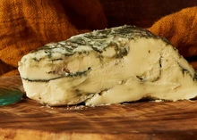 A piece of Brixton Blue, a vegan cheese, reveals blue veining on its yellow interior, highlighting its distinctive appearance and rich flavor