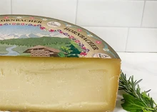 A partially cut wheel of Hornbacher cheese with its lable, rests against white tiled background, accompanied by fresh herbs