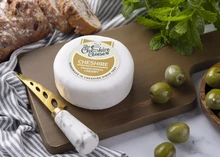 A creamy Cheshire cheese, wrapped in wax and labeled, rests on a wooden board with a knife, slices of bread, olives, and fresh herbs