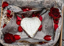 A heart-shaped Clava Brie, adorned with a white bloomy rind, rests in a basket surrounded by red roses and petals