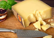 A slice of Comté cheese, along with several chunks, rests on a wooden board, accompanied by a cutting knife and a scattering of fresh herbs.