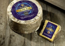 A wheel of Darling Blue cheese with a label sits next to a slice, showing its mold-ripened rind and blue-grey color, on a wooden surface