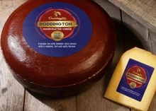 A wheel of Doddington cheese, with its plastic rind, sits next to a slice of the cheese on a wooden surface