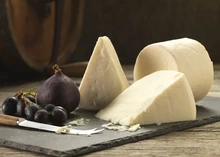 A cylindrical wheel of Elsdon cheese, with large slices, figs, black grapes, and a knife, sits on a black marble slab, all arranged on a wooden surface