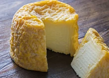 A cylindrical Langres cheese with a partially cut slice reveals its distinctive orangey-brown, wrinkled rind, resting on a wooden surface