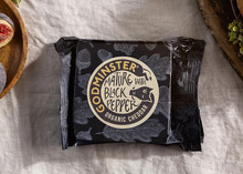 Godminster Mature Bruton Best with Black Pepper Organic Cheddar, wrapped in its paper, rests on a white cloth alongside fresh figs