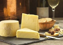 A cylindrical wheel of Northumberland Original Cheese, along with large slices, is arranged on black marble, accompanied by a knife, crackers, and a glass of white wine