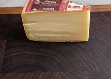 A wedge of wheel-shaped Grand Cru Reserve cheese, with its wrapper revealing a smooth interior, sits on a wooden surface