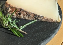 A wedge of Cave Aged Marisa cheese with a dark brown rind rests on a black marble surface, accompanied by fresh herbs.