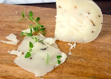 A slice and chunks of Truffle Pecorino (Pecorino al Tartufo) cheese, speckled with delicate truffle flecks, are displayed on a wooden surface, accompanied by fresh herbs
