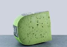 A wheel of Pesto Verde cheese with a slice cut out, showing green interior with basil and garlic flecks