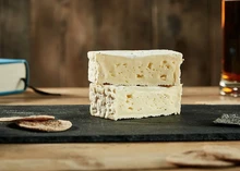 Rectangular pieces of Pont l'Eveque Cheese, stacked to reveal a pale yellow interior, sit on a wooden surface alongside crackers and a glass of beer