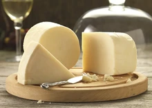 A cylindrical wheel of Redesdale cheese, accompanied by large slices and a knife, rests on a round wooden board, with a glass of white wine visible in the background