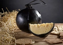 Shorrock's Lancashire Bomb cheese, encased in black wax, with a partially cut piece revealing its smooth interior, is accompanied by crackers and shredded paper, all set on a wooden surface