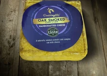 A rectangular block of Smoked Cuddy's Cave Cheese, wrapped in a plastic covering with a blue label, rests on a wooden surface
