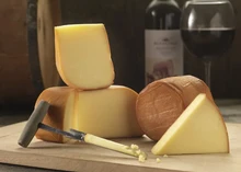 Slices of Northumberland Oak-smoked cheese in various sizes, showcasing their bronzed outer rind, are arranged on a wooden surface alongside a conical cheese sampler, a glass of red wine, and a wine bottle