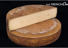 A half-cut wheel of Tomme du Jura cheese sits atop another, both showcasing their orange-colored rind and smooth interior, set against a black background