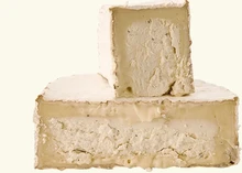 A halved disc of Truffle Tremor cheese with a slice resting on top, revealing its bloomy rind, creamy texture, and flecks of black truffles, set against a white background