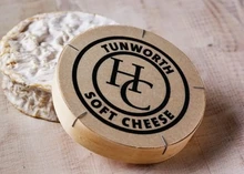 A disc of Tunworth cheese wrapped in paper sits next to its circular box, which displays its name
