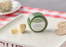 A Yorkshire Wensleydale cheese disc, wrapped in green wax, sits on a white plate alongside crackers, placed on a table