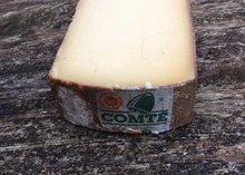 A slice of 'aged Comte 30 months' cheese showcasing its smoth, buttery texture rests on a wooden surface