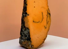 A piece of vegan Balham Blue cheese, showcasing a mold-ripened rind and an orange interior marbled with blue veins, rests on a white surface