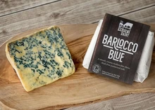 A piece of Blarlocco blue cheese, featuring prominent blue veins, sits on a wooden board beside another wrapped Blarlocco blue cheese