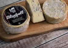 A disc of Bluebell cheese with a label, another without, and a half-cut piece revealing blue veining and a smooth interior, resting on a wooden board