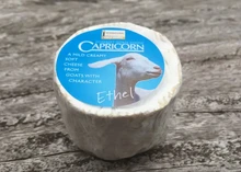 A cylindrical-shaped Capricorn Somerset Goat's Cheese, wrapped in a transparent sheet with a blue label, rests on a wooden surface