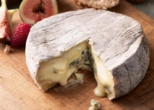A disc of Cote Hill Blue cheese, partially sliced to reveal its creamy, runny interior with blue veins, rests on a wooden surface next to fresh figs