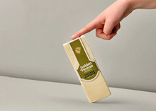 A rectangular-shaped Golden Hooves Vintage Cheddar, in its wrapper, is held by the tip of a finger against a beige background