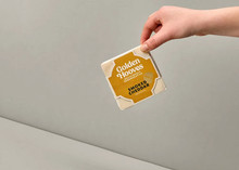 A square-shaped Golden Hooves Smoked Cheddar, in its wrapper, is held in a hand against a beige background