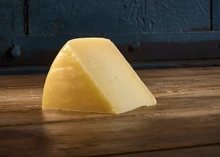 A triangular piece of Pecorino cheese rests on a wooden surface and dark blue color wall in the background.