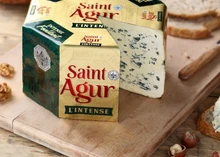 A partially cut wheel of Saint Agur cheese with its wrapper, set on a wooden board alongside a knife and some nuts