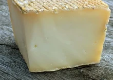A slice of Sheep Rustler cheese, highlighting its distinctive orange-brown rind, rests on a wooden surface