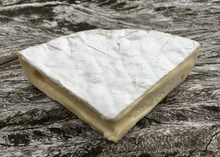 A triangular slice of Cornish Brie cheese, revealing its smooth, buttery interior, sits on a wooden board