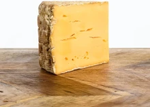 A piece of Woodside Red cheese rests on a wooden revealing orange interior and a natural rind.
